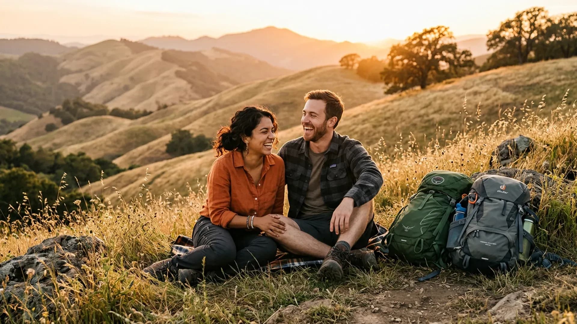 Young couple laughing naturally on a hillside at sunset with hiking backpacks